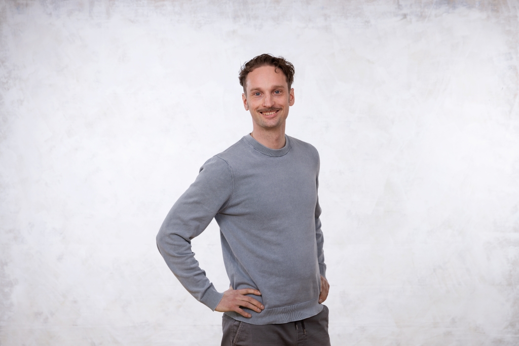 Rabbe Sandström has his hands on his waist, he looks into the camera and smiles. Background is a grey canvas.