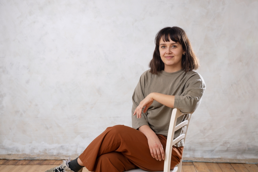 Siiri Halko sits in a chair, looks into the camera and smiles. The background is a grey canvas.