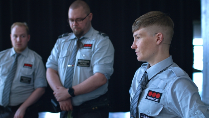 Kolme henkilöä vartijan asussa. Kaksi näistä seisoo taaempana ja he näkyvät epätarkkana. Etummainen henkilöistä on taiteilija Pilvi Takala, johon kuva on tarkentunut.