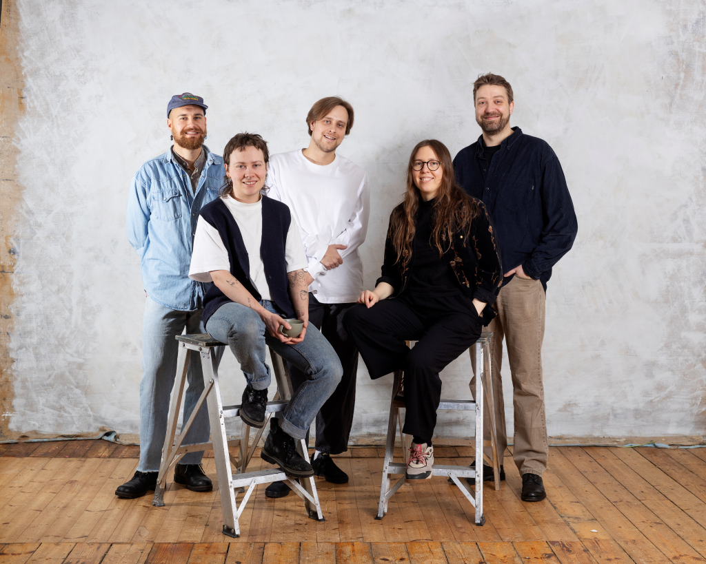 The filmmakers are in the centre of the picture, they look at the camera and smile. Two are sitting on low ladders in the foreground, the other three are standing behind them. In the background there is a wooden floor and a light-coloured wall.