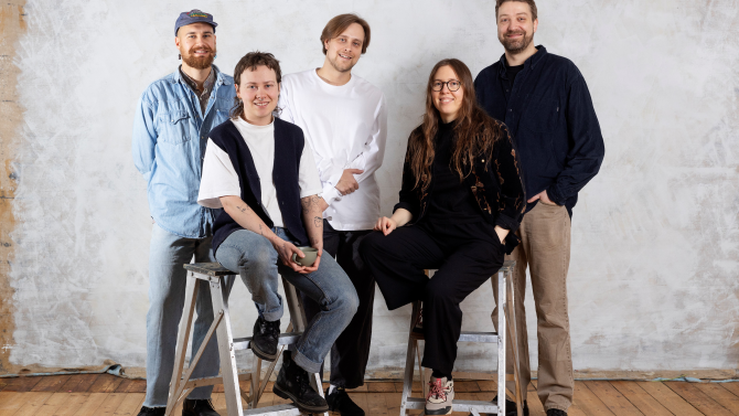 The filmmakers are in the centre of the picture, they look at the camera and smile. Two are sitting on low ladders in the foreground, the other three are standing behind them. In the background there is a wooden floor and a light-coloured wall.