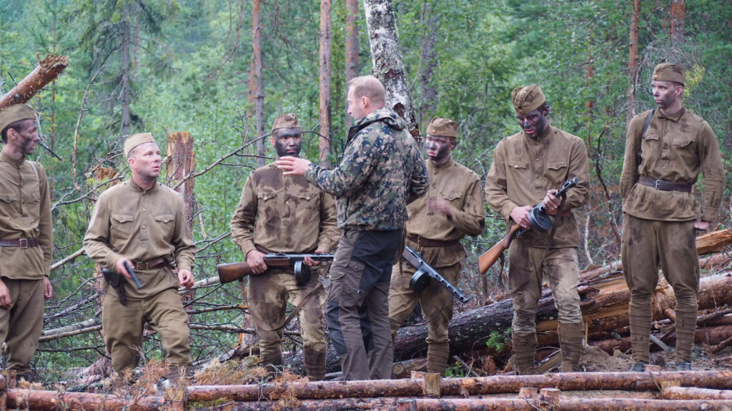 Asevastaava Kari Lehtinen on kuvassa keskellä ja opastaa joukkoa näyttelijöitä Tuntemattoman sotilaan kuvauksissa metsässä. 