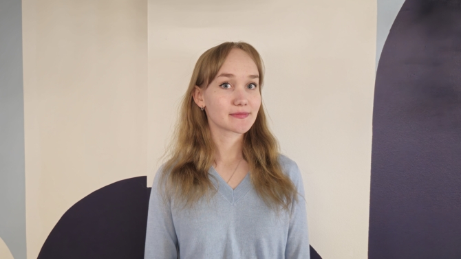 Emma Koponen katsoo kameraan ja hymyilee. Taustalla seinä, jossa on sinisiä kuvioita.