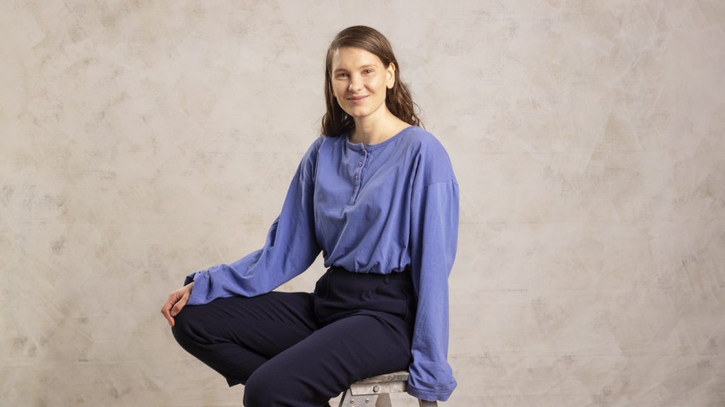Laura Rantanen istuu tuolilla, toinen käsi on polvella. Hän katsoo kameraan ja hymyilee. Tausta on vaalea.