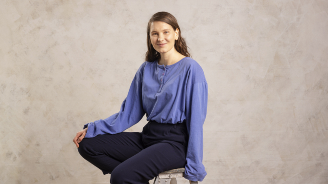 Laura Rantanen istuu tuolilla, toinen käsi on polvella. Hän katsoo kameraan ja hymyilee. Tausta on vaalea.